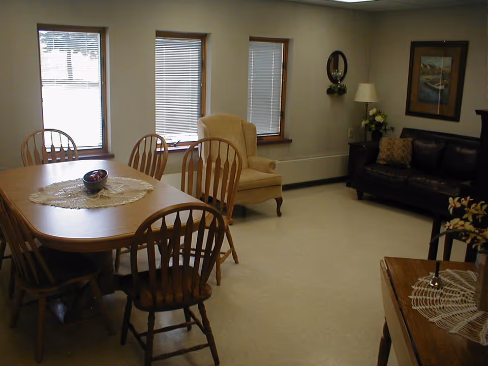 Communal dining and sitting area with a wooden dining table and chairs, an armchair and a sofa by windows.