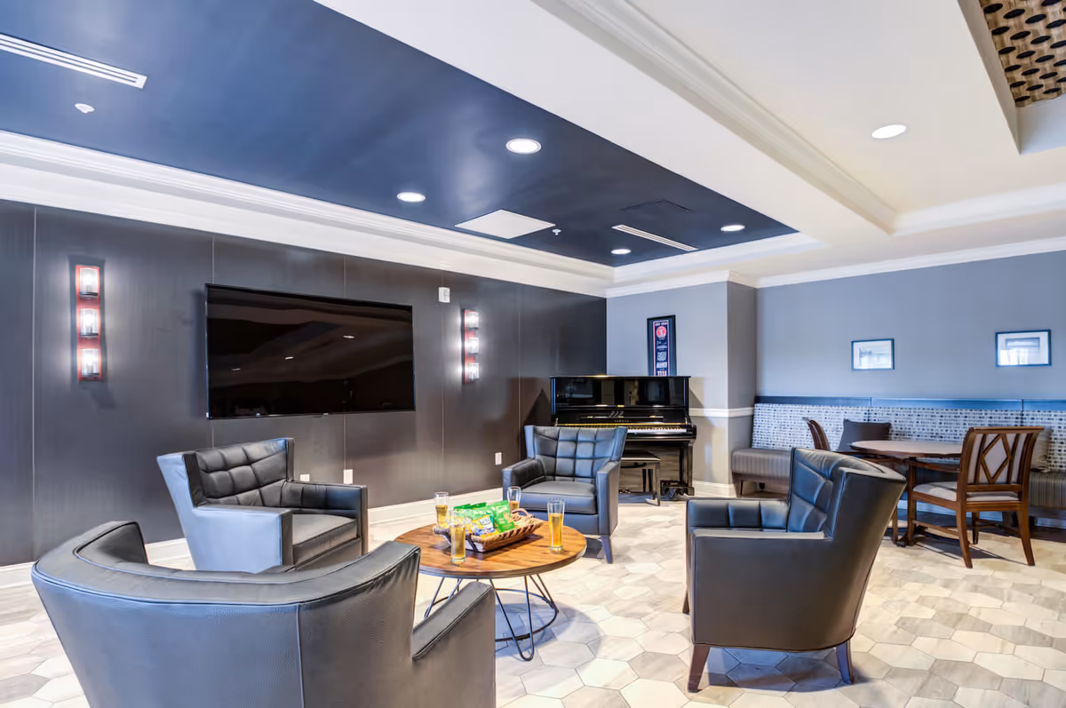 A modern common lounge with black leather armchairs around a round wooden coffee table, a wall-mounted TV, and an upright piano in the background.