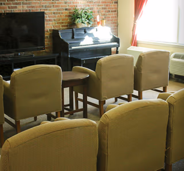 A cozy living room area with several beige upholstered chairs arranged facing a flat-screen TV and a black upright piano against a brick wall. A small round wooden table is placed between some of the chairs. Sunlight streams in through a window with red curtains on the right side.