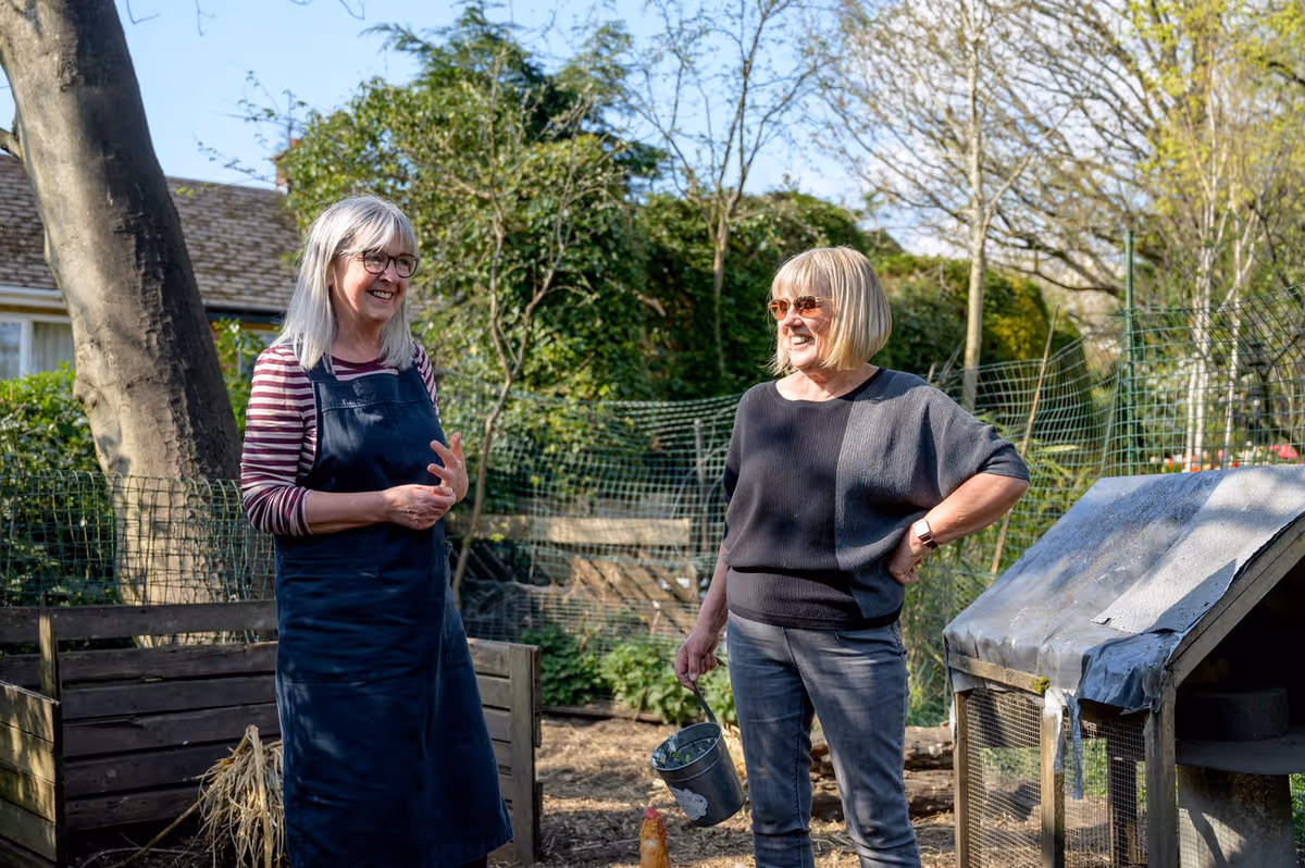 Two older women standing outdoors in a garden area, smiling and talking. One woman is wearing a striped long-sleeve shirt with a dark apron, and the other is wearing sunglasses and a dark sweater, holding a small bucket. There is a wooden compost bin and a small animal coop nearby, with trees and greenery in the background.