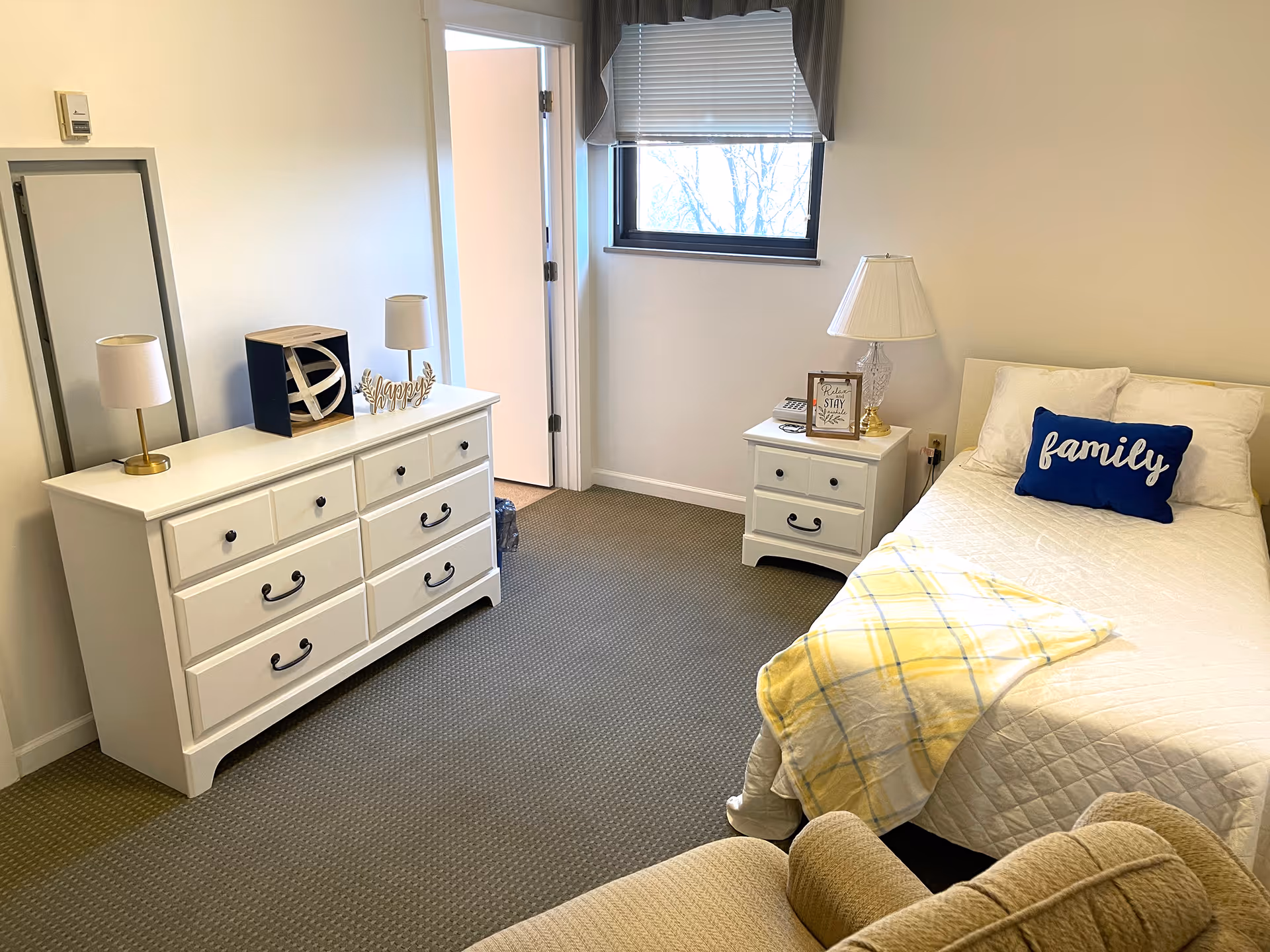 A cozy bedroom with a white dresser topped with two lamps and decorative items, a window with a blind partially drawn, a white nightstand with a lamp and framed quote, a bed with white bedding and a blue pillow labeled 'family', and a yellow plaid blanket draped over the bed. A beige armchair is partially visible in the foreground.