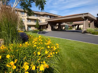Exterior view of a multi-story senior living facility with a covered entrance driveway. The foreground features a well-maintained lawn and vibrant yellow flowers under a clear blue sky.