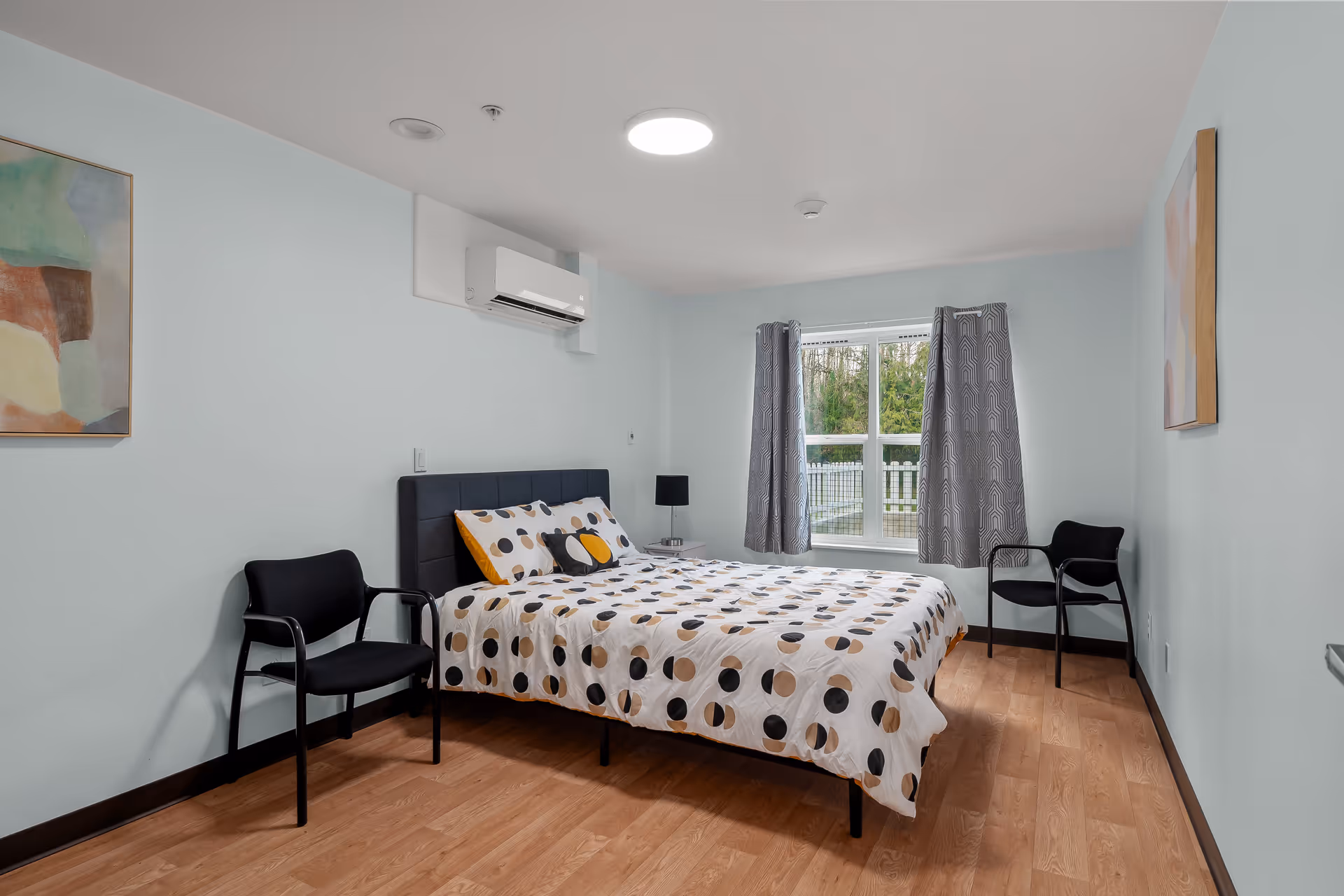 A simple bedroom with a double bed dressed in polka-dot bedding, two chairs, a window with curtains, and light wood flooring.