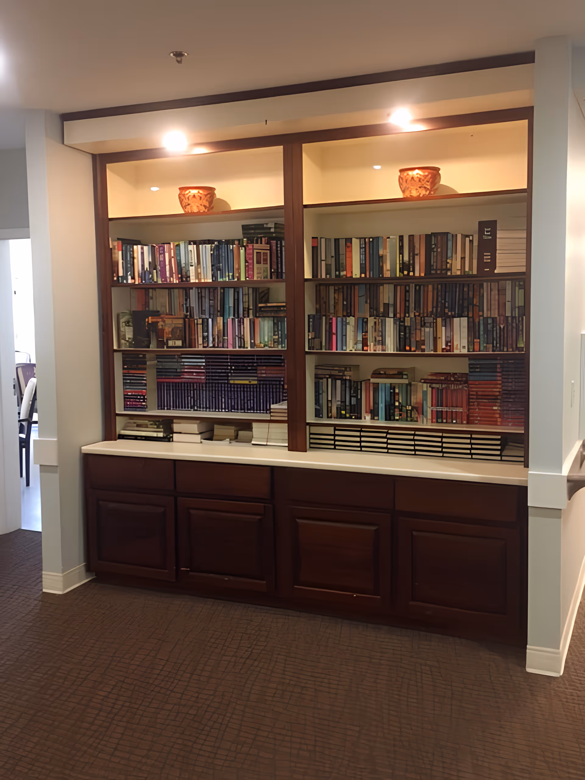 A built-in wooden bookshelf with multiple shelves filled with books and two decorative pots on the top shelf, illuminated by overhead lights. The bookshelf is set against a wall with a white countertop and dark wooden cabinets below. The floor has a textured brown carpet, and there is a doorway visible to the left leading to another room with chairs and tables.