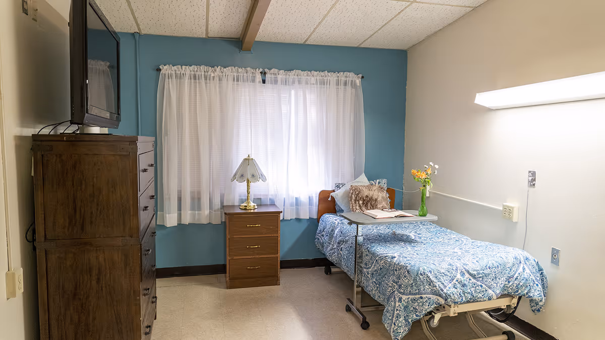 A small furnished bedroom with a single hospital-style bed, bedside table and lamp, wooden dresser with TV, and a curtained window.