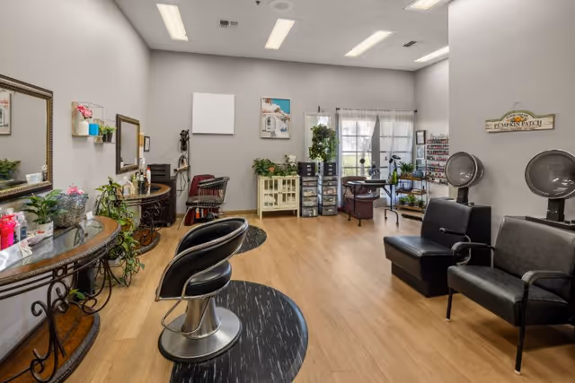 Interior view of a senior living community salon with two black salon chairs in front of mirrors on the left, two black hair dryer chairs on the right, wooden flooring, plants, and various salon equipment and supplies arranged around the room.