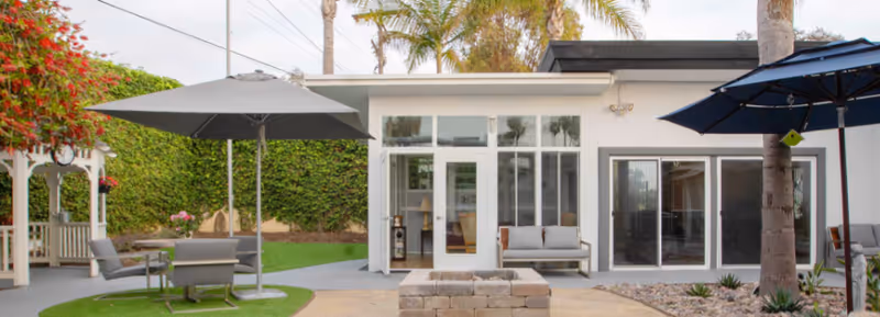 Outdoor patio area at Avantgarde Senior Living | la Jolla featuring a white building with large windows and sliding glass doors, two large umbrellas shading seating areas with chairs and tables, a fire pit made of bricks in the center, palm trees, and a green hedge in the background.