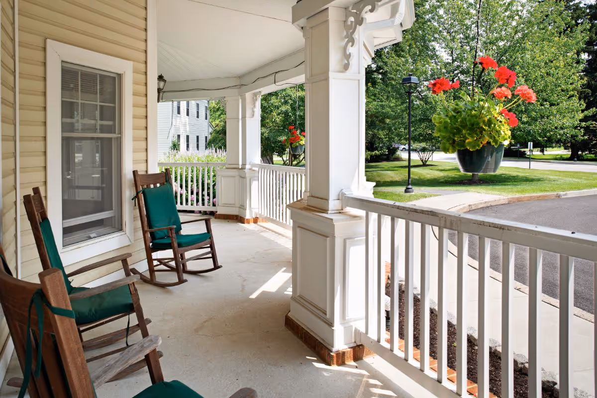 A covered porch area with white railings and columns, featuring three wooden rocking chairs with green cushions. A hanging flower pot with red flowers is visible, along with a view of a paved driveway and green trees in the background.