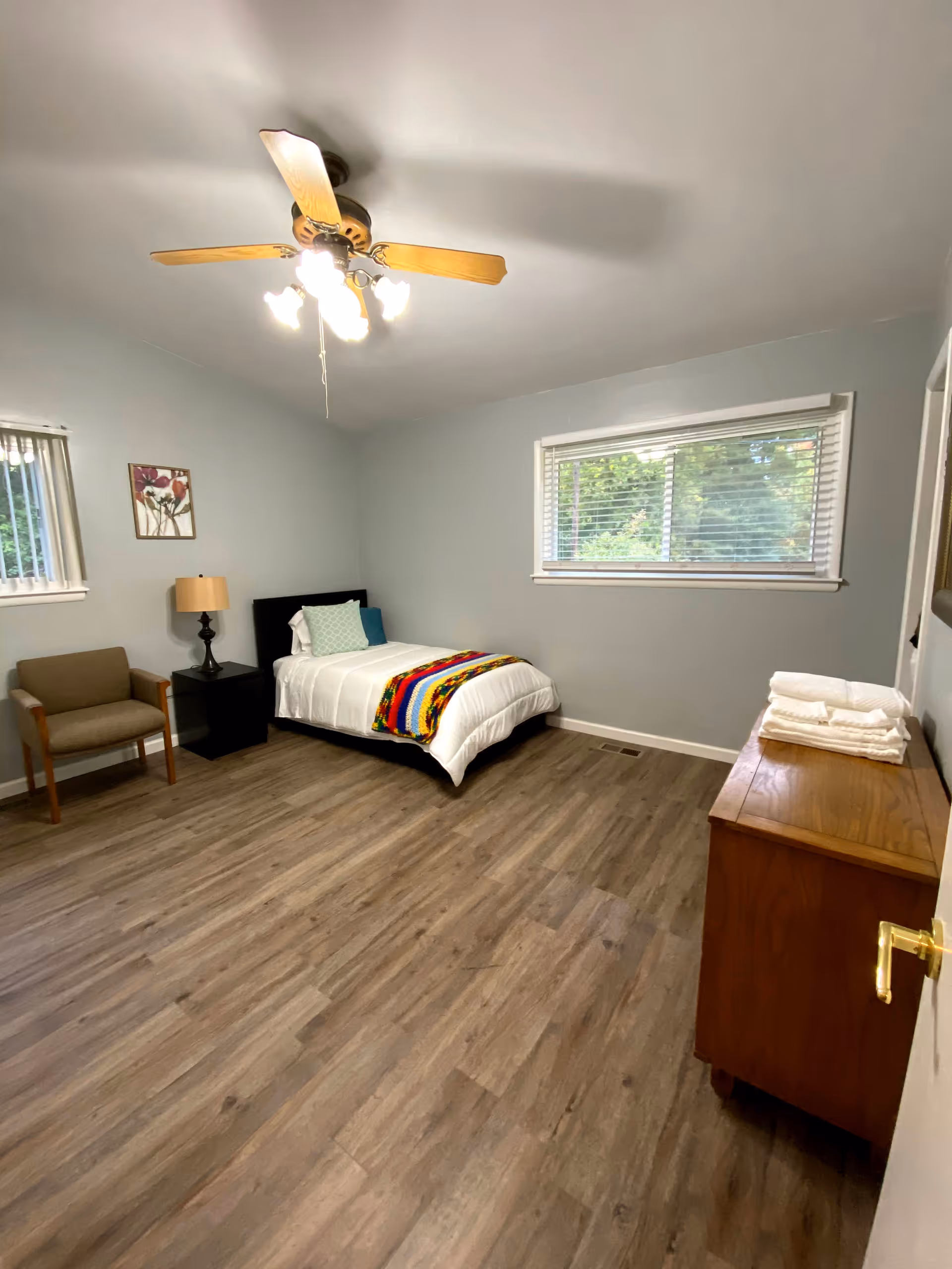 A simple bedroom with a single bed covered with white bedding and a colorful blanket at the foot. There is a wooden chair with armrests, a black nightstand with a lamp, a wooden dresser with folded towels on top, and two windows with blinds letting in natural light. The room has light gray walls, wood flooring, and a ceiling fan with lights.