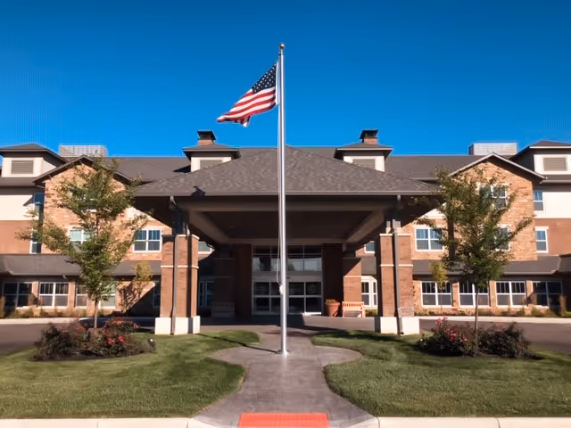 Front entrance of a multi-story brick retirement residence with a covered portico, landscaped lawn, and an American flag on a flagpole.