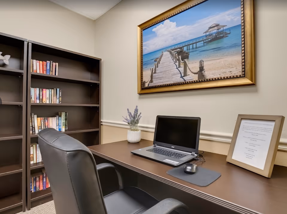 A small office or study area with a dark wooden desk and black leather chair. On the desk is a laptop, a mouse on a mousepad, a small potted plant, and a framed document. Behind the desk is a dark wooden bookshelf with several books. Above the desk hangs a large framed picture of a wooden pier extending into a blue ocean under a partly cloudy sky.