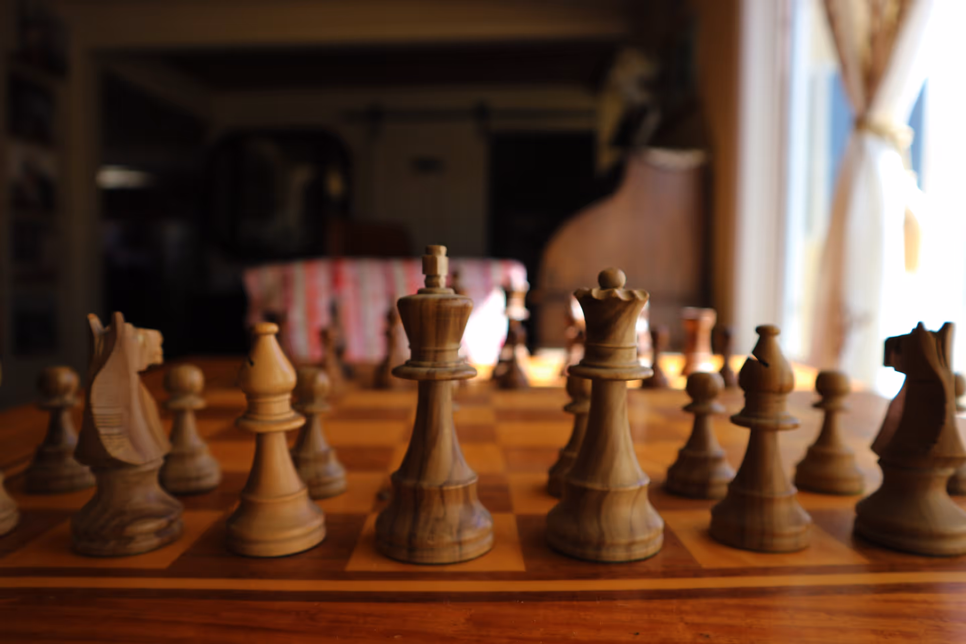 Close-up view of a wooden chessboard with chess pieces arranged for a game, set on a wooden table in a warmly lit room with a window and curtains in the background.