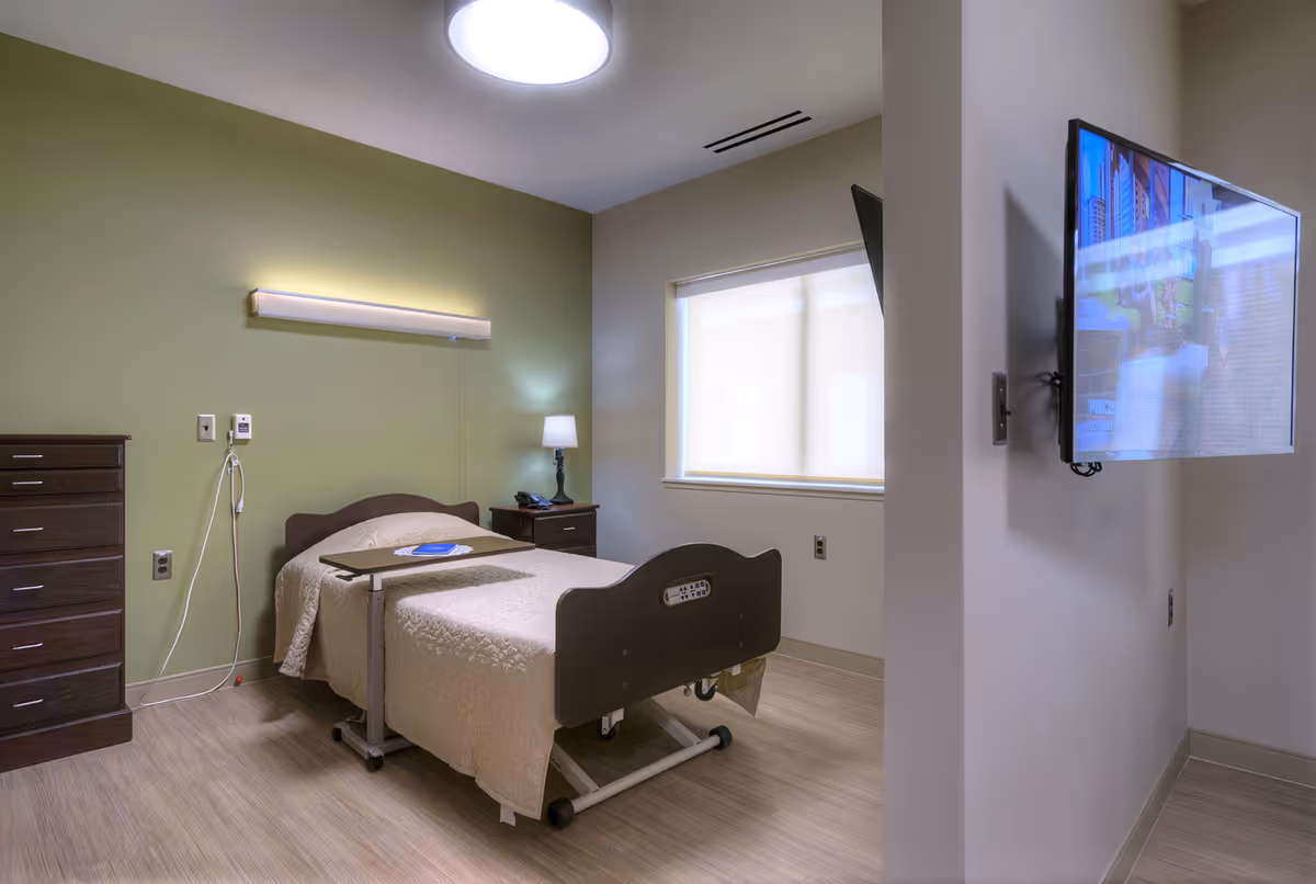 A simple, clean bedroom in a retirement community featuring a single hospital-style bed with a beige quilt, a dark wooden dresser and nightstand, a lamp, a wall-mounted light above the bed, and a flat-screen TV mounted on a partial wall. The room has light-colored walls with one green accent wall and a window with a closed blind.