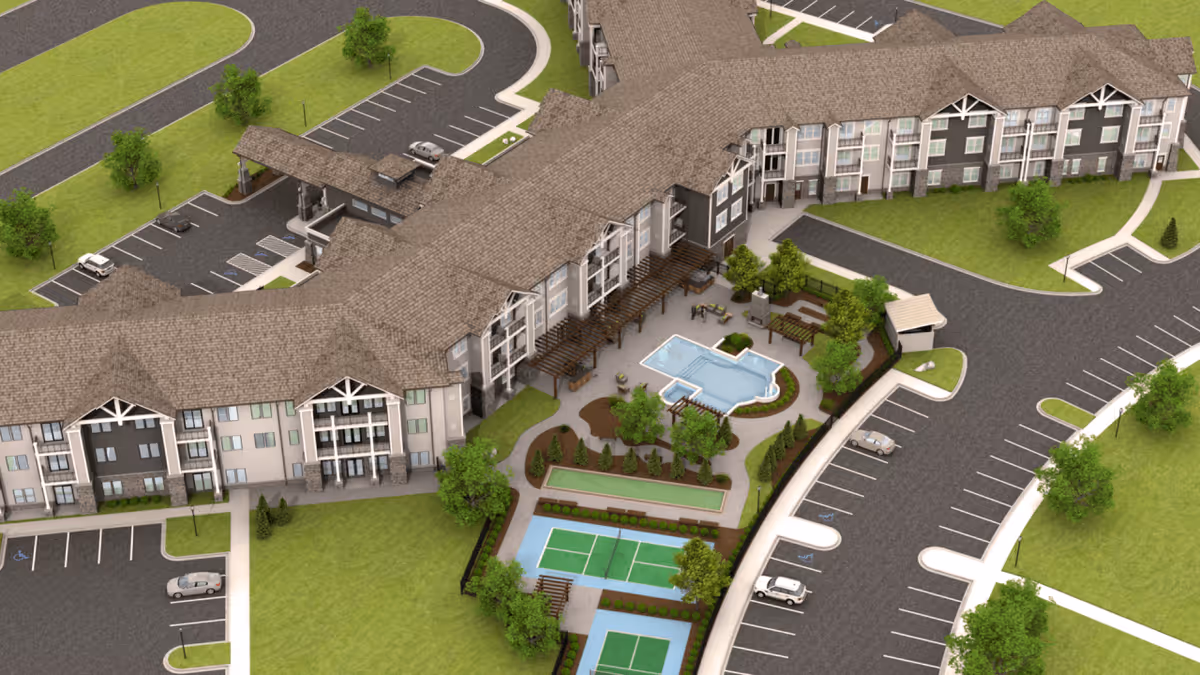 Aerial view of Monark Grove Madison senior living facility showing a large multi-story building with a brown roof, surrounding parking lots, green lawns, trees, and outdoor amenities including a swimming pool, seating areas, and two tennis or pickleball courts.