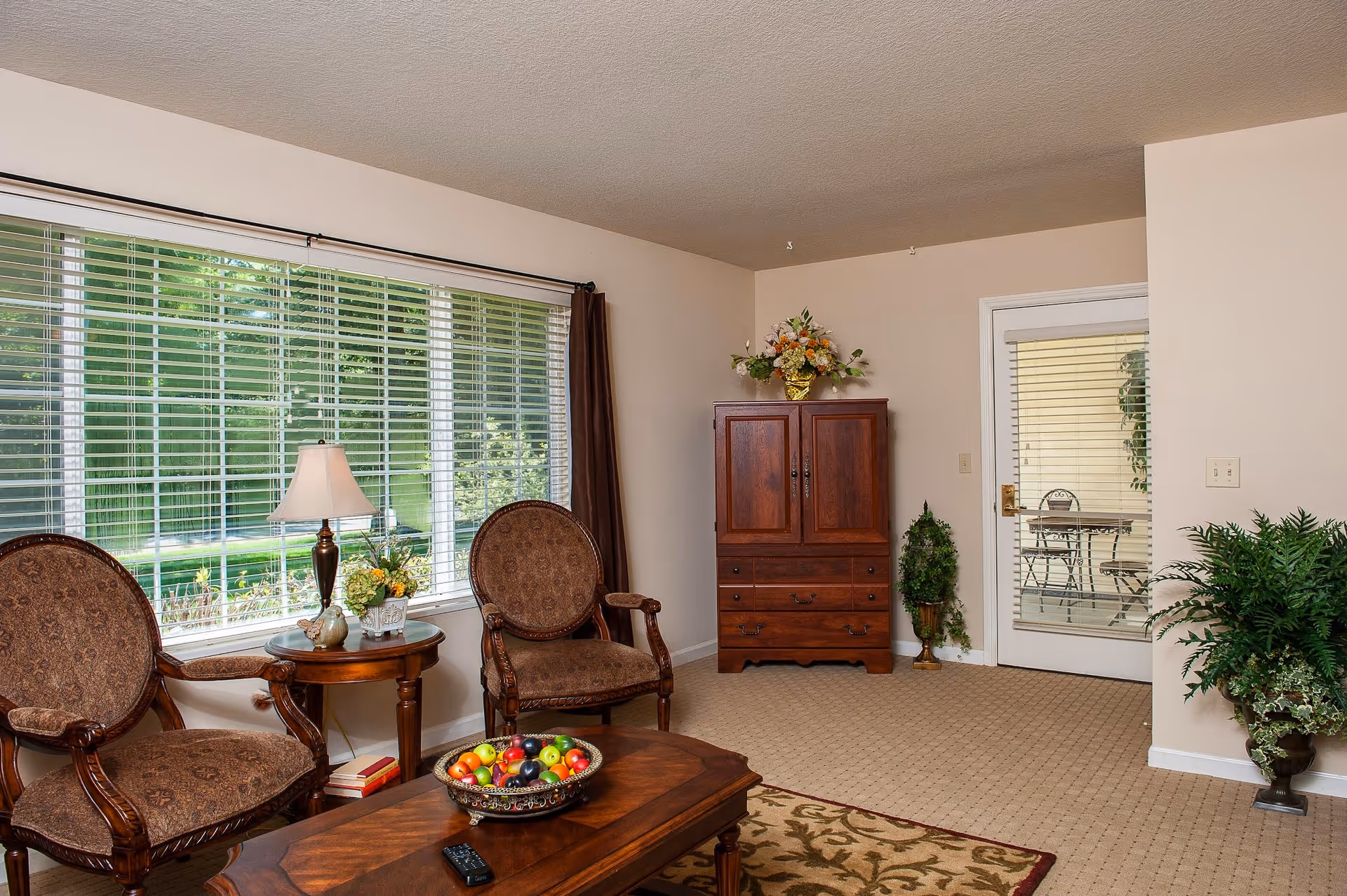 A cozy living room area with two upholstered armchairs, a wooden coffee table with a decorative bowl of fruit and a remote control, a side table with a lamp and flower arrangement, a wooden cabinet with a floral arrangement on top, and a glass door leading to an outdoor patio. The room has beige walls, carpeted floor, and large windows with white blinds letting in natural light.