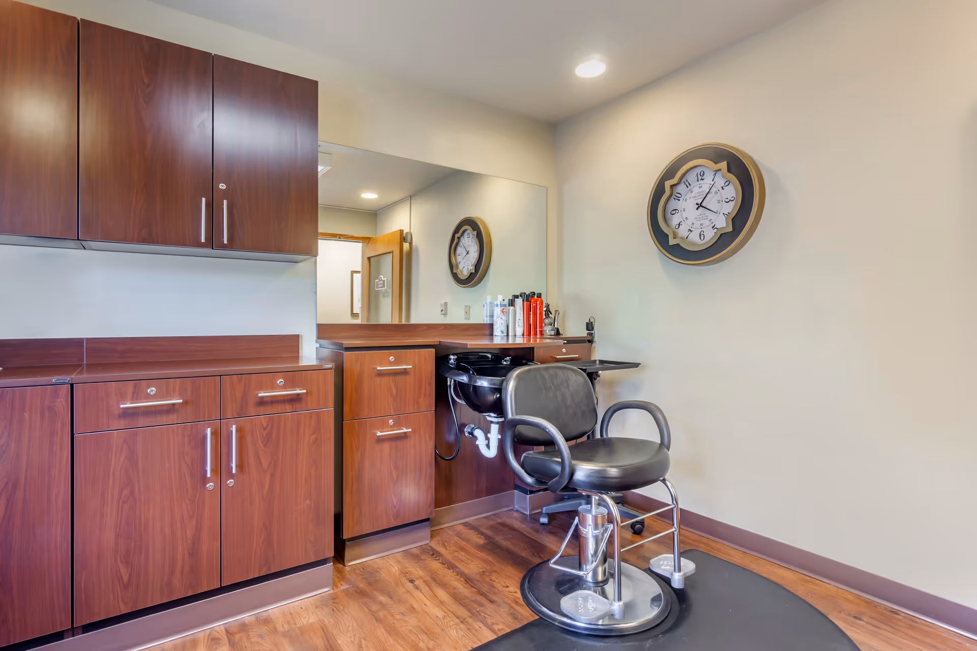 Interior of a hair salon area in a senior living facility with wooden cabinets, a large mirror, a black salon chair, a black hair washing sink, and a decorative wall clock showing the time as 12:10.