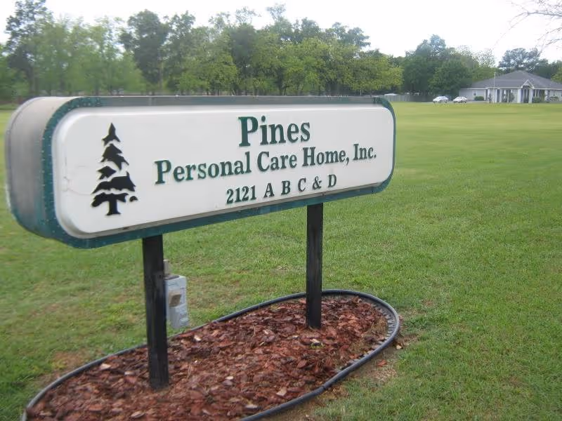 A sign for Pines Personal Care Home, Inc. with the address 2121 A B C & D, placed on a grassy area with trees and a building visible in the background.