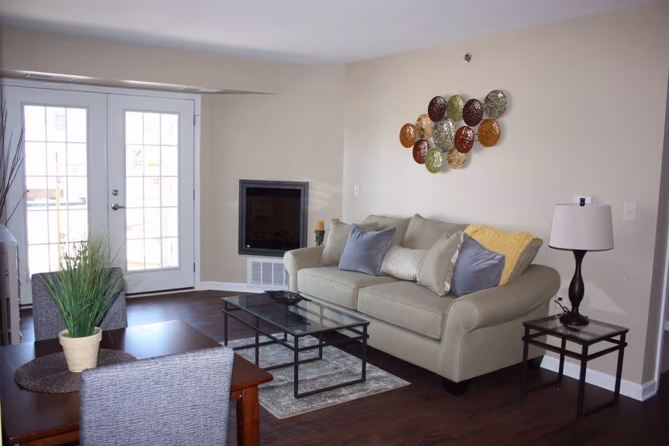 A cozy living room with a beige sofa adorned with multiple cushions, a glass coffee table on a small rug, a side table with a lamp, and a decorative wall art piece above the sofa. There is a dining table with chairs and a potted plant in the foreground. French doors with glass panes lead outside, allowing natural light to fill the room.