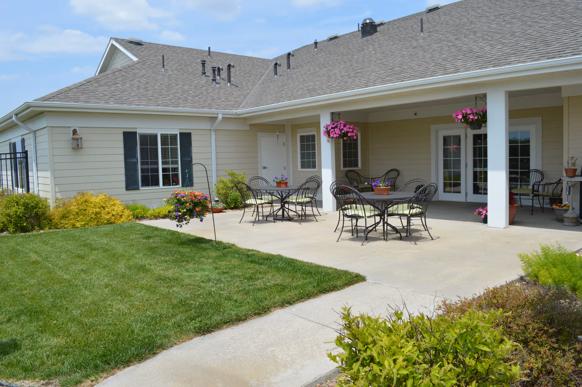 Outdoor patio area of a senior living facility with several black metal tables and chairs with green cushions. Hanging flower baskets and potted plants decorate the space. The patio is adjacent to a beige building with white trim and a gray roof. There is a well-maintained lawn and garden surrounding the patio.