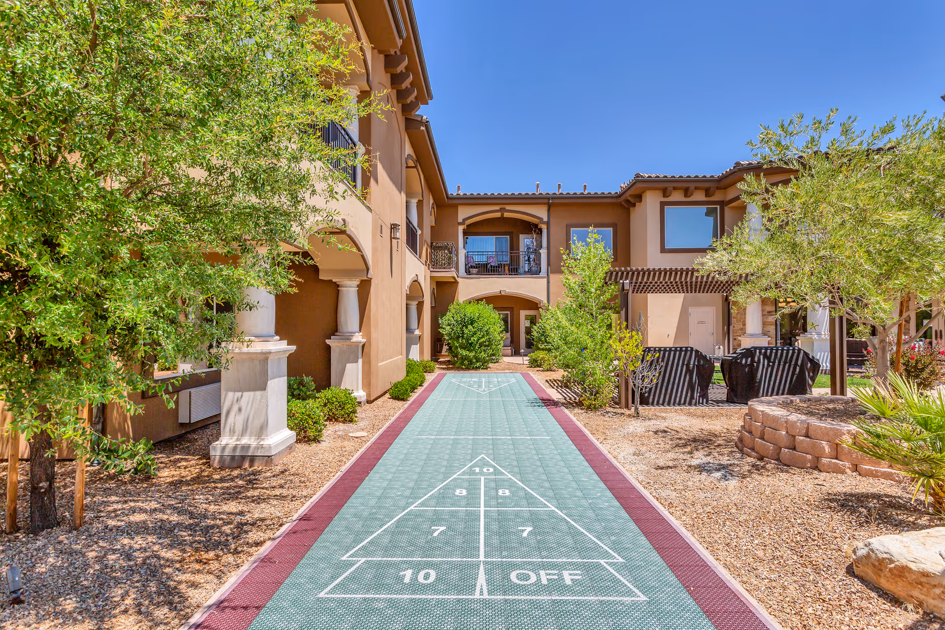 Outdoor courtyard area of a senior living facility with a shuffleboard court in the center, surrounded by trees, shrubs, and a two-story building with balconies and columns under a clear blue sky.