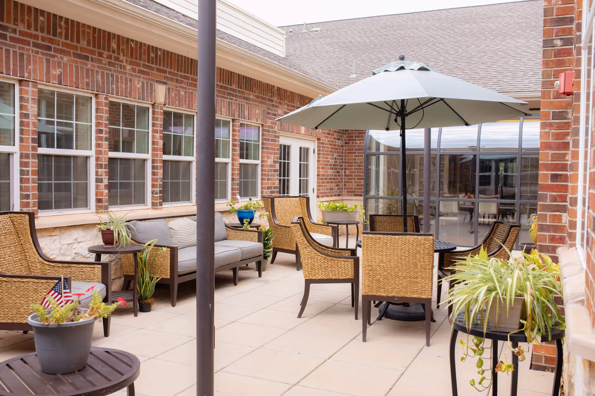 Outdoor patio area with wicker chairs, a round table with a large umbrella, potted plants, and a brick building with multiple windows in the background.