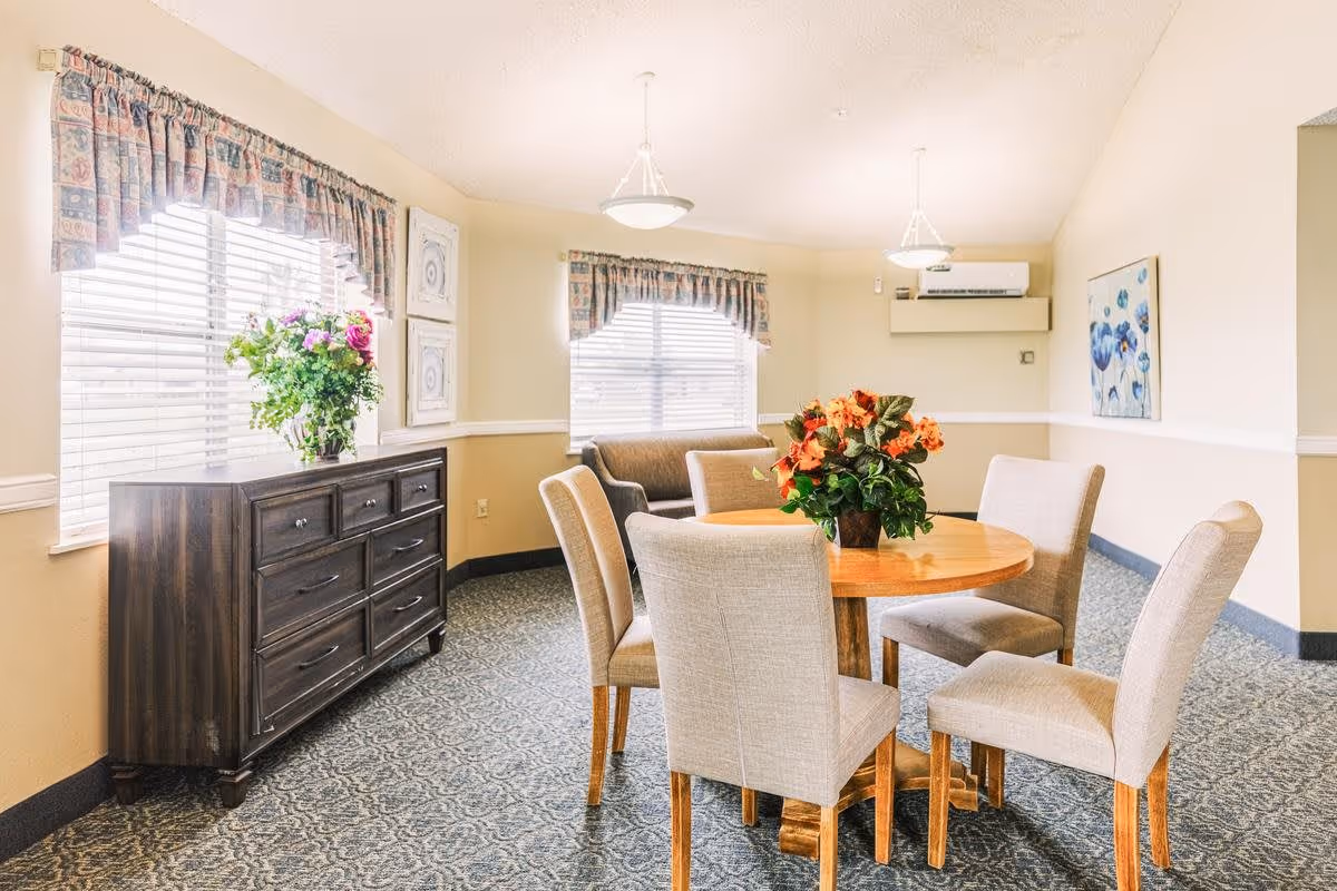 A bright room with a round wooden table surrounded by six beige upholstered chairs. There is a floral centerpiece on the table. The room has two windows with patterned valances, a dark wooden sideboard with a vase of flowers on top, a small sofa, and a wall-mounted air conditioning unit. The walls are painted light beige with a white chair rail, and a framed floral artwork hangs on one wall.