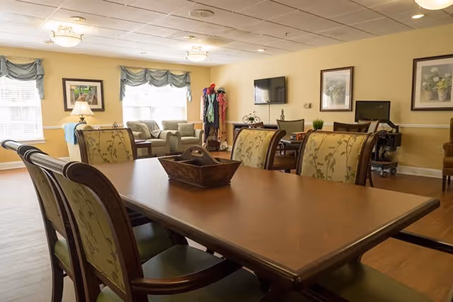 A well-lit common area in a senior living facility featuring a large wooden table with six cushioned chairs around it. In the background, there are comfortable armchairs, a wall-mounted TV, framed artwork, and windows with green valances allowing natural light to fill the room.