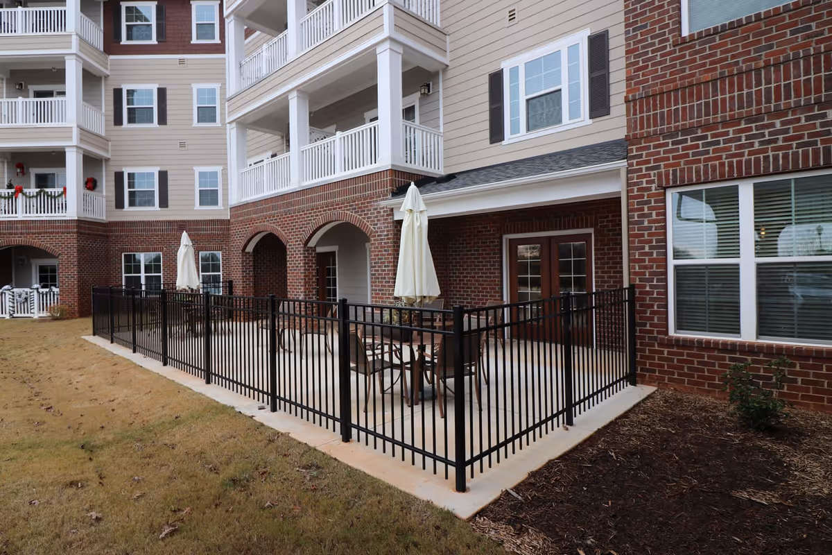Outdoor patio area at a senior living facility with a black metal fence surrounding a concrete patio. The patio has a table with chairs and a closed white umbrella. The building exterior features red brick on the lower level and beige siding with white trim on the upper levels, along with balconies and windows.