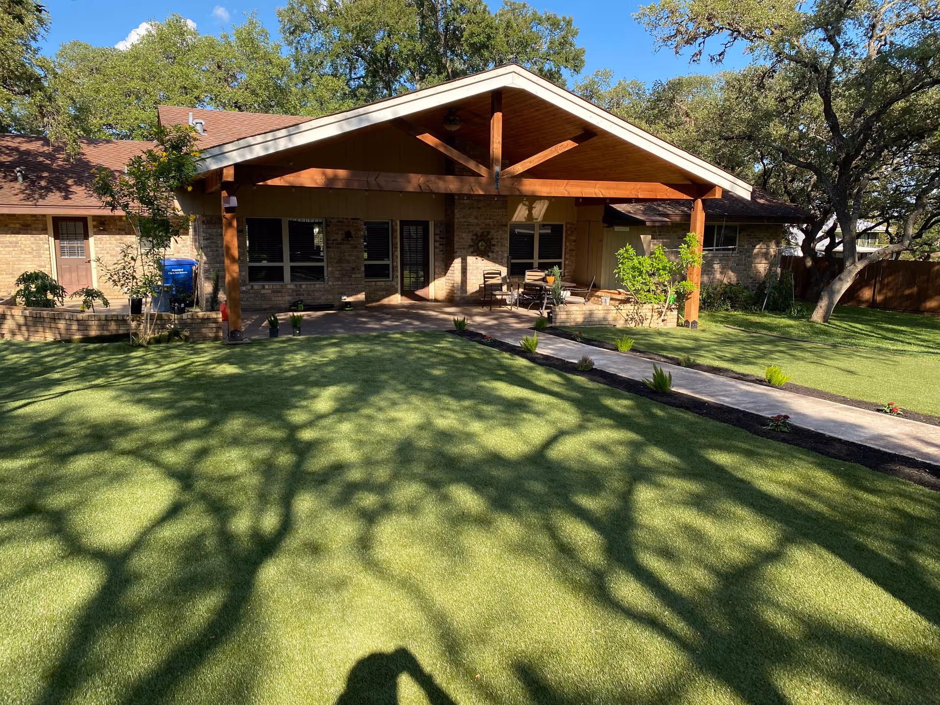 Single-story brick building with a wooden covered patio, outdoor seating, and a manicured lawn with large tree shadows.