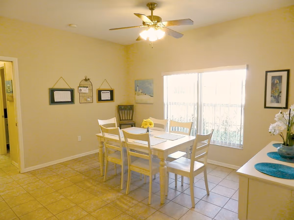 A bright dining room with a white rectangular table and six matching chairs. The table has placemats and a small vase with yellow flowers. There is a ceiling fan with lights above the table. The room has tiled flooring and light-colored walls with framed pictures and decorations. A large window with vertical blinds lets in natural light. A white sideboard with blue placemats and a vase with white flowers is on the right side.