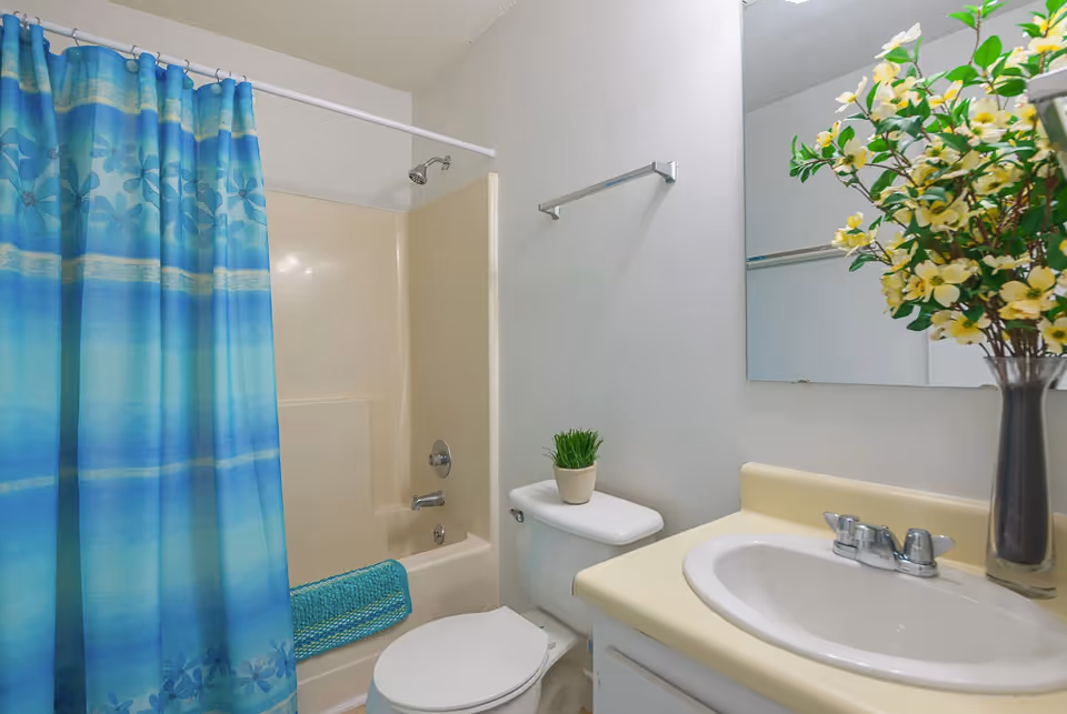 Bathroom with a blue patterned shower curtain, bathtub/shower, toilet with a small potted plant, sink with mirror, and a vase of yellow flowers on the counter.