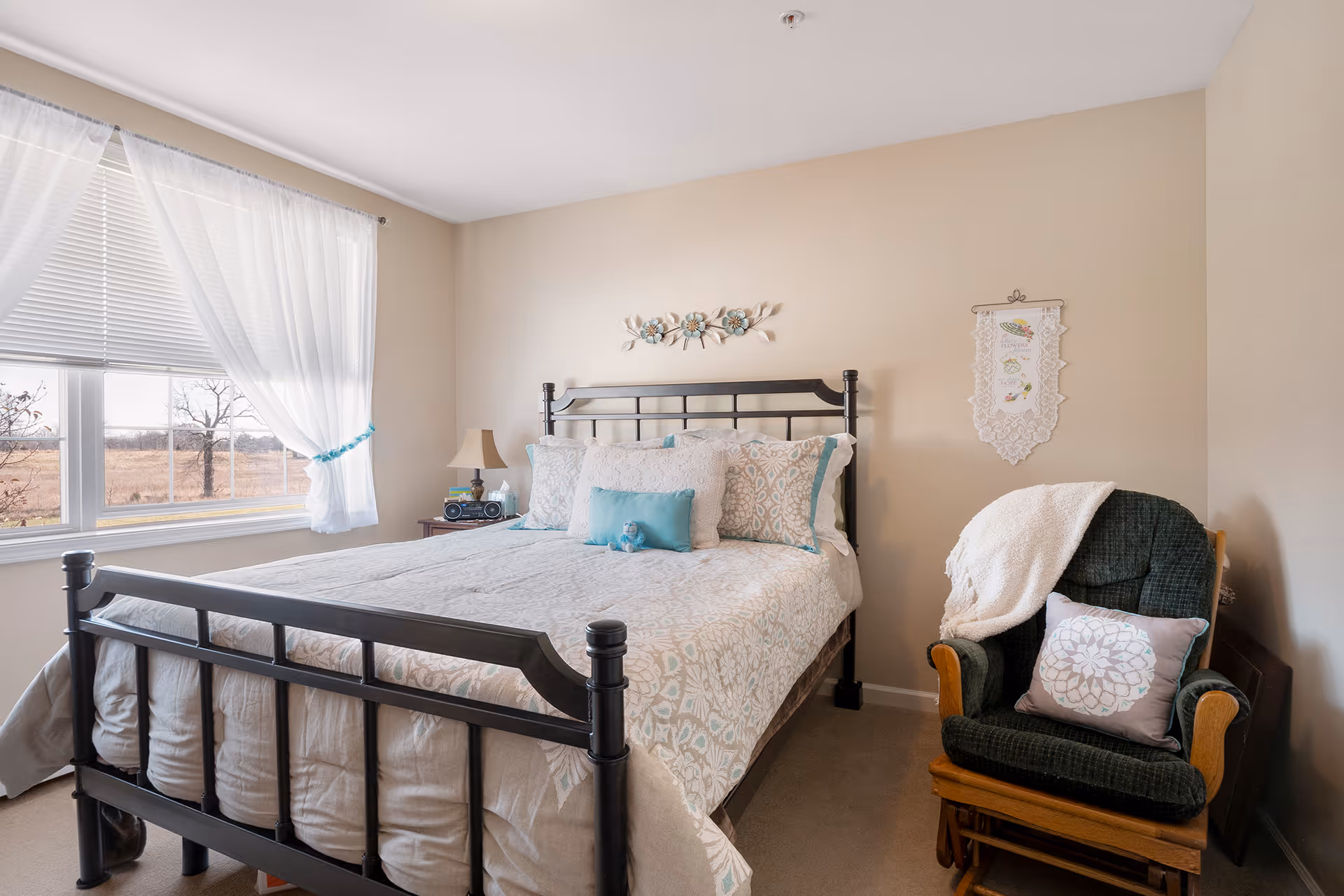 Sunlit bedroom featuring a black metal-framed bed with patterned bedding, a bedside table under a window with sheer curtains, and a cushioned armchair.