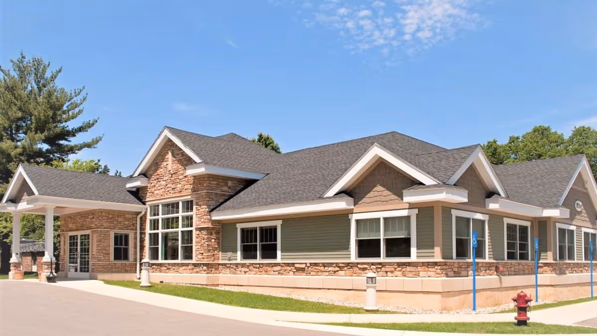 Exterior view of a single-story nursing and rehabilitation center building with stone and siding facade, multiple windows, a covered entrance, and a clear blue sky above.
