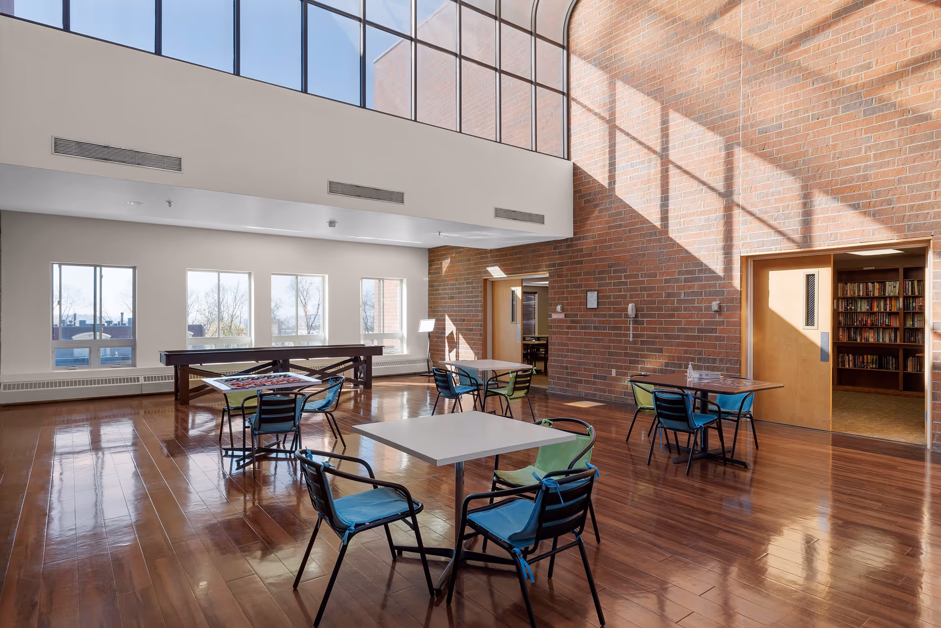 A bright, spacious common area with high ceilings and large windows letting in natural light. The room features polished wooden floors, several tables with colorful chairs, and a shuffleboard table near the windows. A doorway leads to a room with bookshelves filled with books.