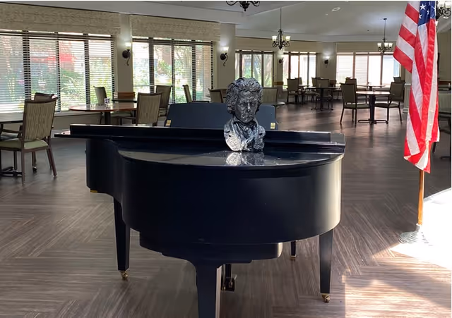 Black grand piano with a bust on top in a spacious dining area with tables, chairs, large windows, and an American flag.