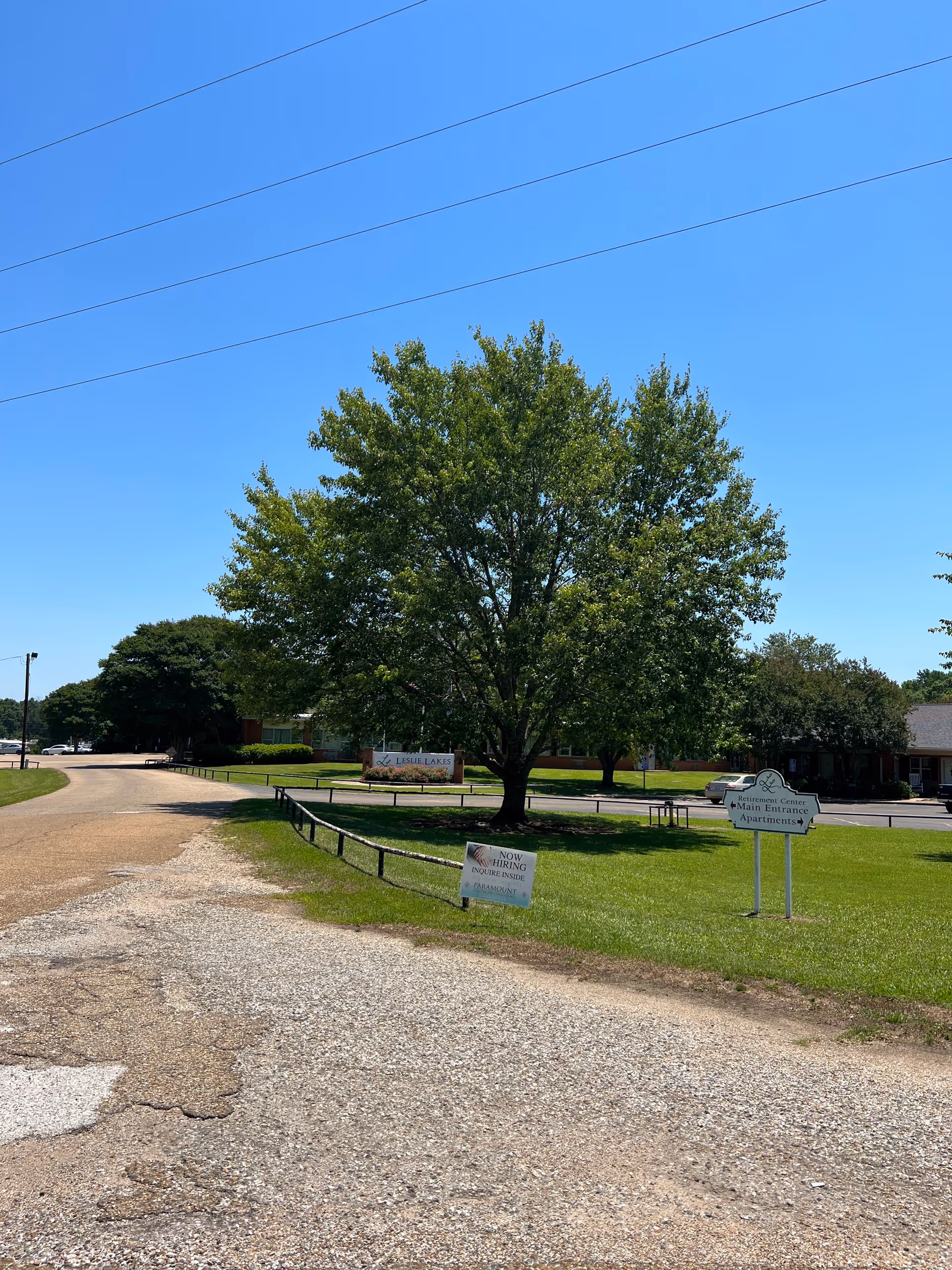 Entrance of Leslie Lakes Retirement Center showing a large tree on a grassy lawn, facility signs, and a gravel driveway under a clear blue sky.