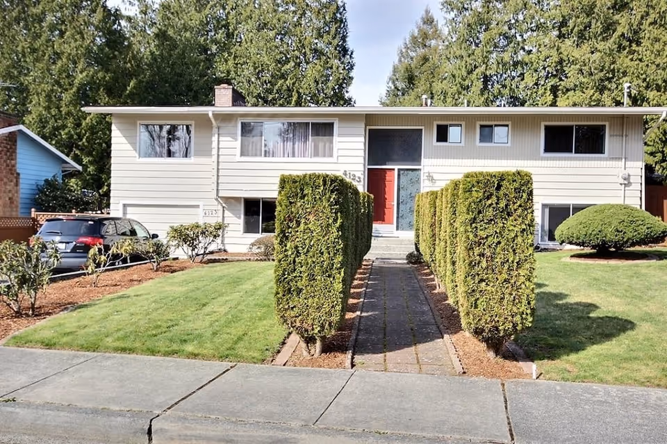 Front exterior view of a two-story residential building with a red front door, large windows, and a pathway lined with neatly trimmed bushes leading to the entrance. The lawn is well-maintained with green grass and some small shrubs. A car is parked on the left side of the house.