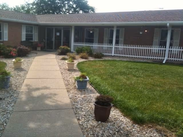 Front exterior view of a single-story brick building with a covered porch, white railing, and a concrete walkway leading to the entrance. The walkway is bordered by small potted plants and landscaping with grass and white gravel.