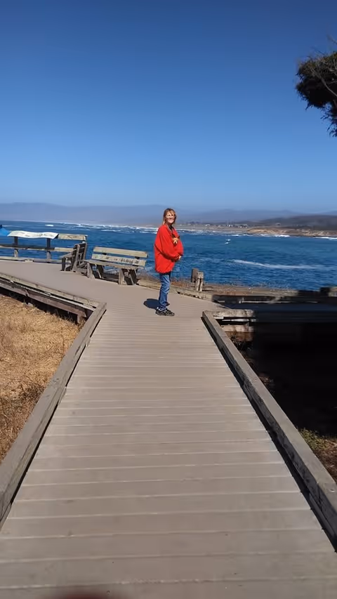 A person wearing a red jacket and blue jeans standing on a wooden boardwalk by the ocean with benches along the side and a clear blue sky overhead.