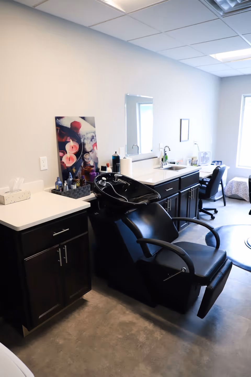 Interior view of a hair washing station in a senior living facility, featuring a black reclining salon chair with a black sink basin attached. The room has dark wood cabinets with white countertops, various hair care products, a mirror, and a sink. In the background, there is a desk with a chair and a window letting in natural light.