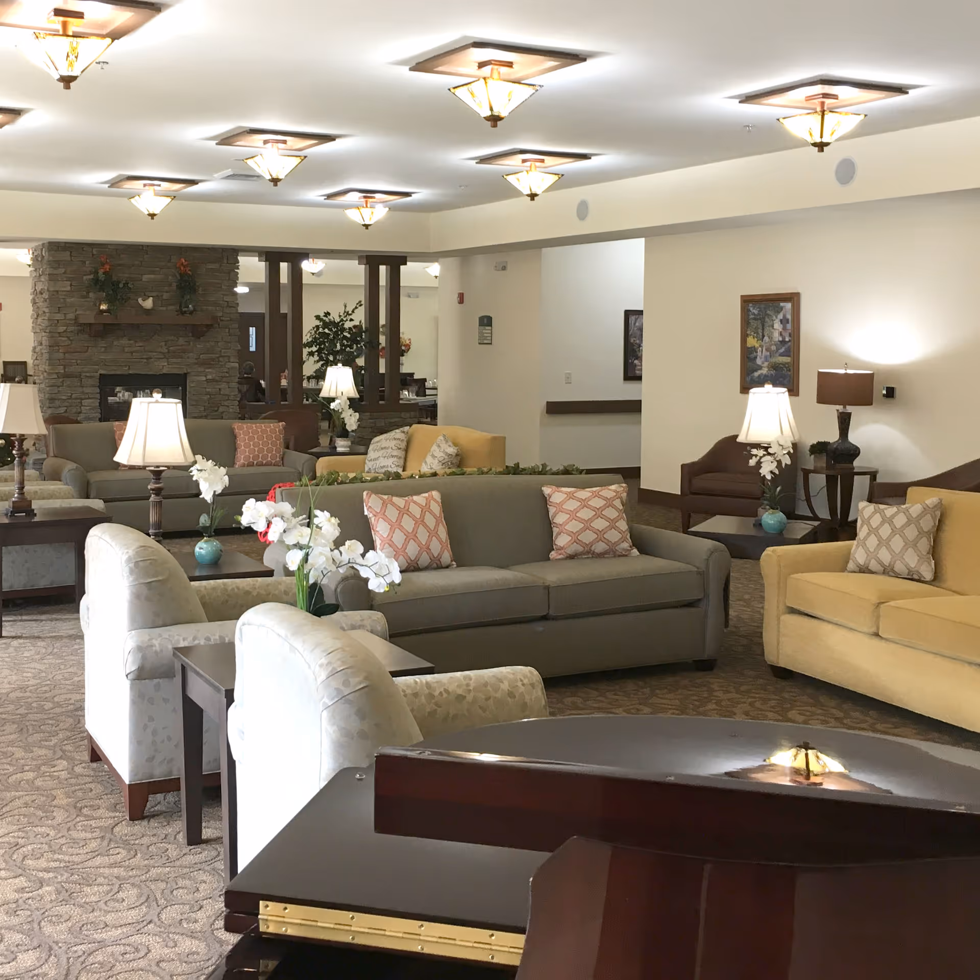 A spacious and well-lit senior living facility common area with multiple sofas and armchairs arranged around coffee tables. The room features a stone fireplace, decorative lamps, floral arrangements, framed artwork on the walls, and ceiling lights with wooden accents.