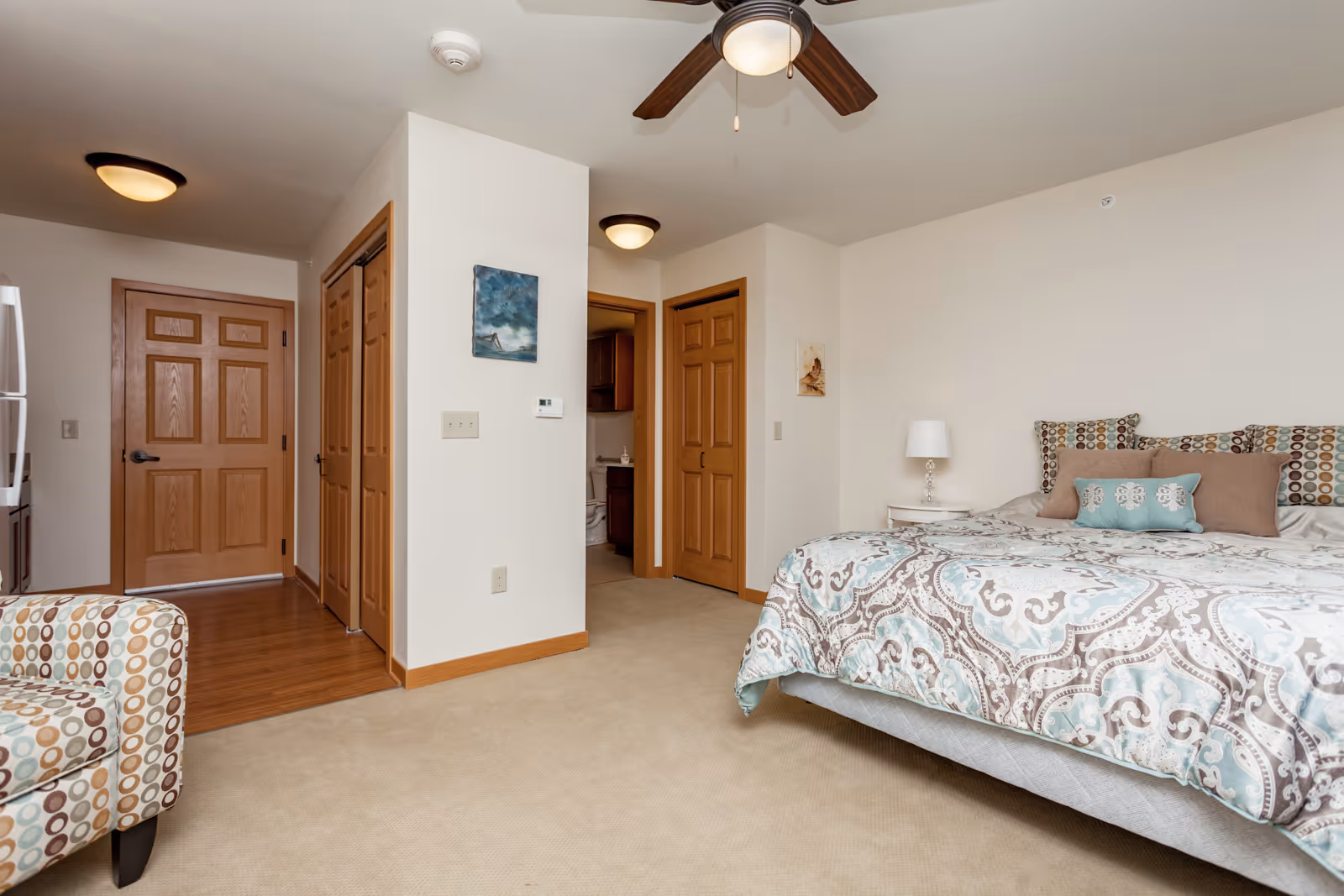 A bedroom in a senior living facility with a large bed covered in a patterned comforter and multiple pillows. There is a small white nightstand with a lamp next to the bed. The room has beige carpet and cream-colored walls with wooden trim. A ceiling fan with a light is mounted on the ceiling. To the left, there is a patterned armchair and a hallway leading to a wooden door and a bathroom with visible toilet and sink.