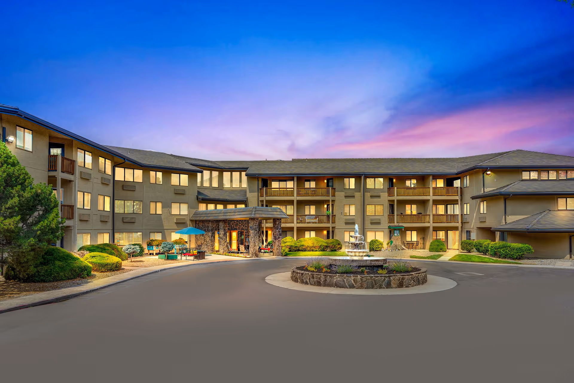 Exterior view of a multi-story senior living facility building at dusk with warm interior lights visible through the windows. The building surrounds a circular driveway with a stone fountain in the center, landscaped bushes, and outdoor seating with umbrellas near the entrance.