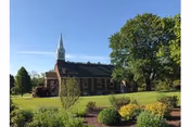 A brick building with a white steeple surrounded by green grass, trees, and landscaped bushes under a clear blue sky.