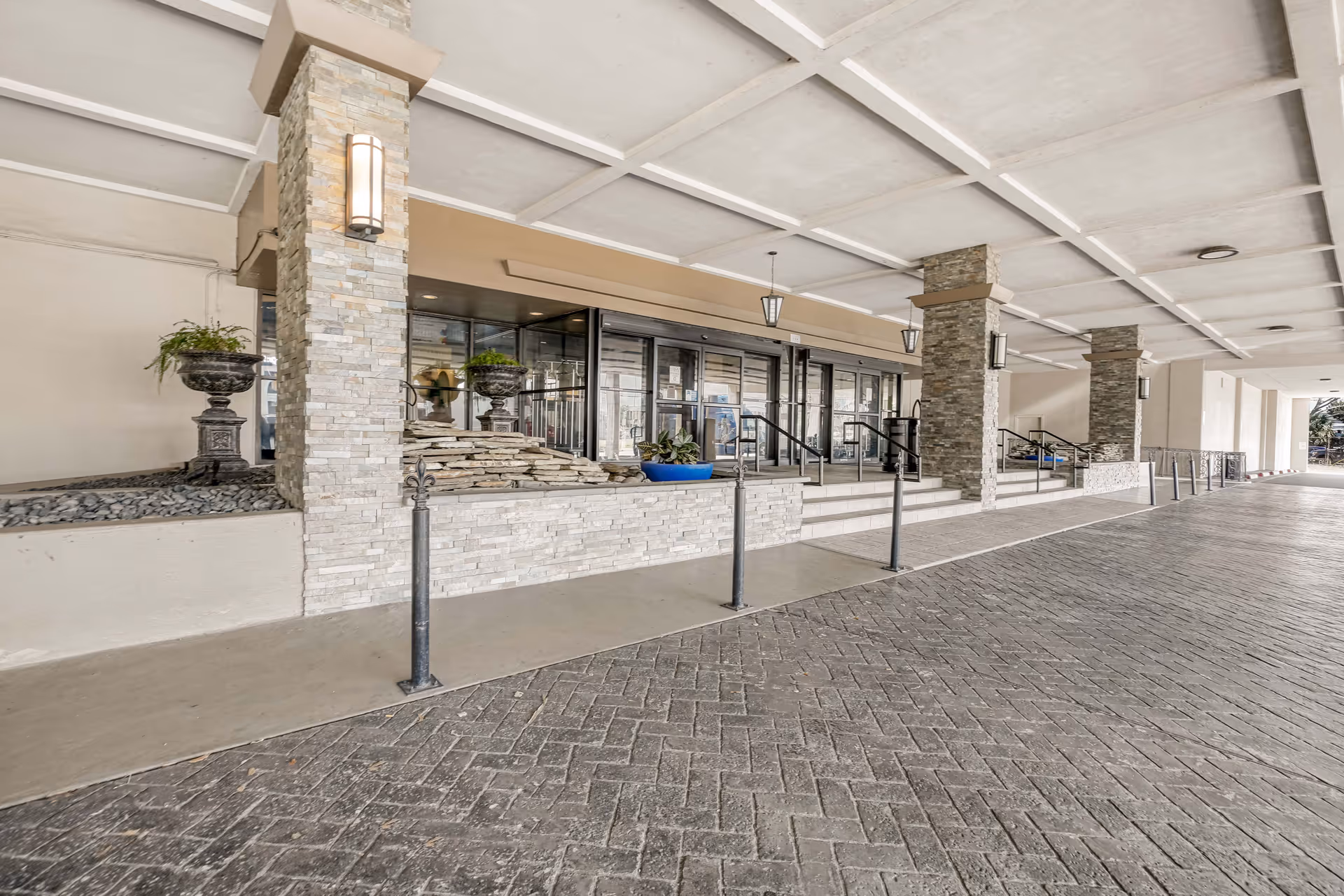 Covered entrance area of a building with stone pillars, steps leading to glass doors, decorative planters, and a paved driveway.