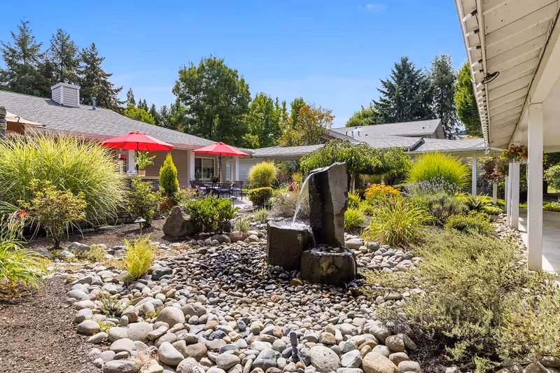 Outdoor garden area at a senior living facility featuring a stone water fountain surrounded by rocks and various green plants. In the background, there are buildings with gray roofs, red patio umbrellas, and outdoor seating. The sky is clear and blue.