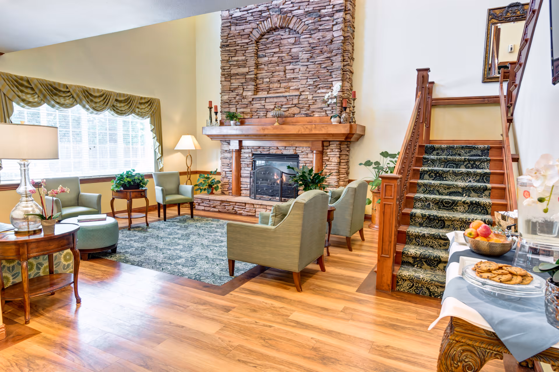 Cozy communal living room with armchairs around a stone fireplace, a patterned rug, wooden staircase, and a refreshment table.