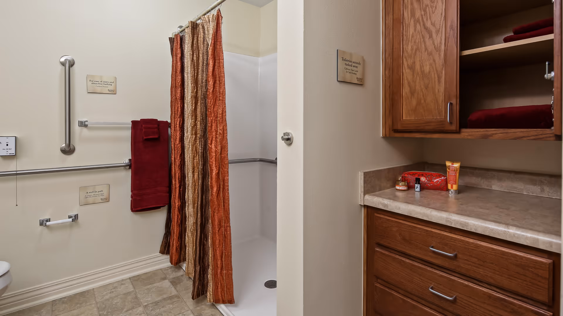 A bathroom with a walk-in shower featuring a multi-colored shower curtain in shades of orange and brown. There are grab bars on the walls for safety, a red towel hanging on a towel rack, and a toilet paper holder. To the right, there is a countertop with wooden cabinets above and below, holding some toiletries and a folded red towel inside the cabinet.