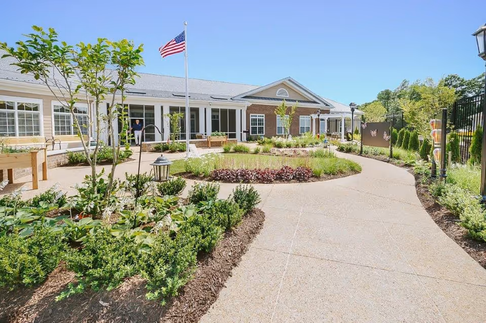 Sunlit landscaped courtyard with a winding walkway, garden beds, and the entrance of a one-story senior living building with an American flag.