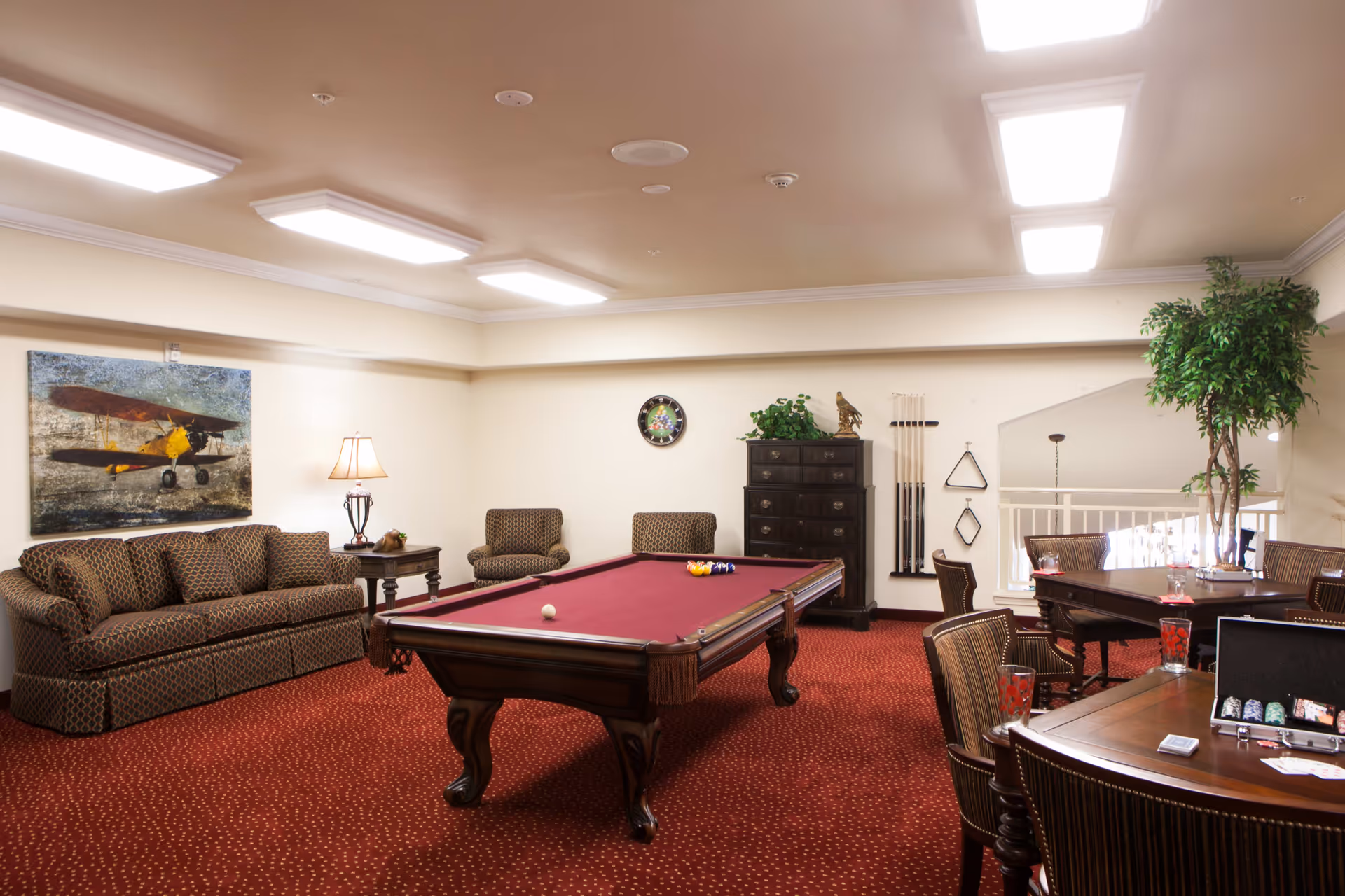 A spacious recreational room with a red carpet and beige walls. The room features a pool table with a red felt surface in the center, a patterned sofa and armchairs along one wall, a side table with a lamp, a large painting of a vintage airplane, a tall chest of drawers with a decorative eagle statue and plants on top, a rack holding pool cues and triangle racks, and a card table with chairs and poker chips. A large potted plant is in the corner near a railing overlooking a lower level.
